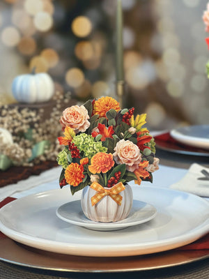 Orange and yellow fall bouquet in a white pumpkin pop card on a dinner plate.