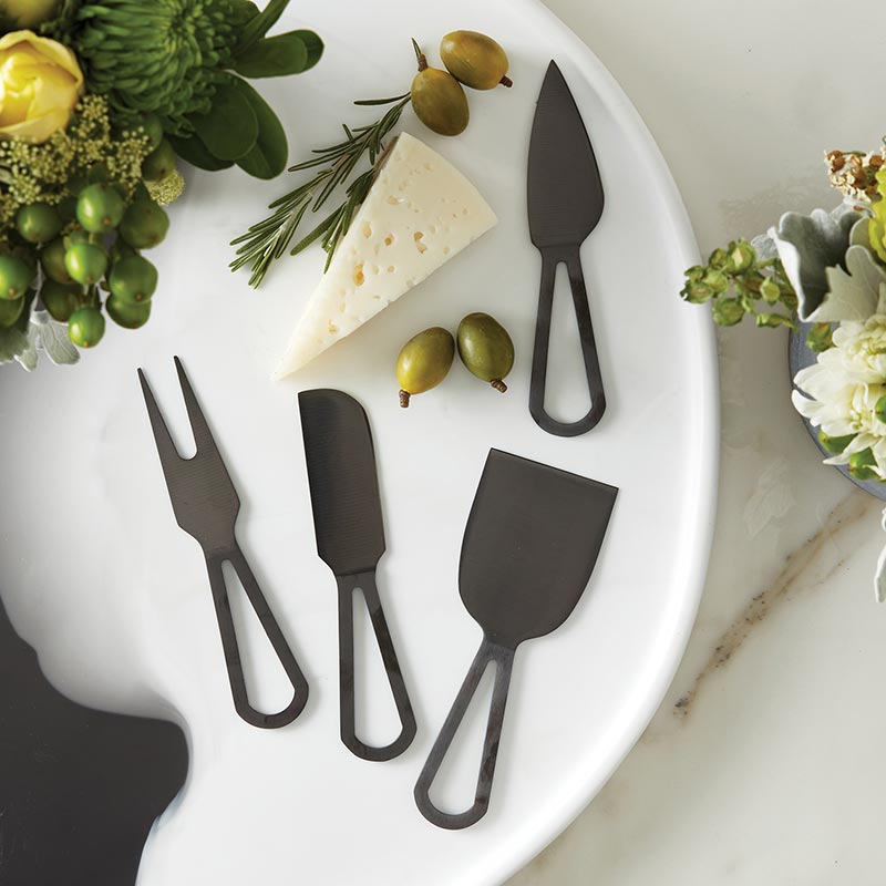 Four matte black cheese knives on a marble round with cheese and olives.