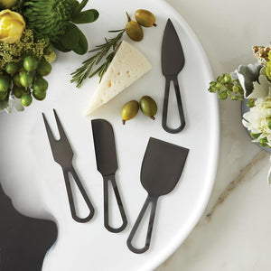 Four matte black cheese knives on a marble round with cheese and olives.