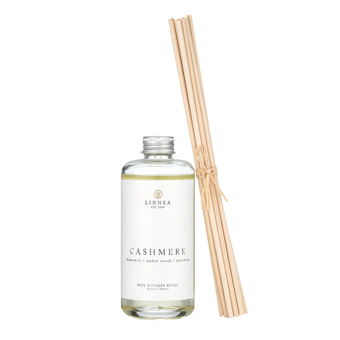 Glass bottle with white label of diffuser refill scent, with ten tied rattan reeds leaning against it.