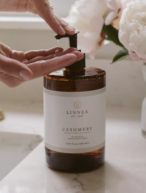 Person pumping liquid hand soap out of an amber Linnea dispenser with white peonies nearby.