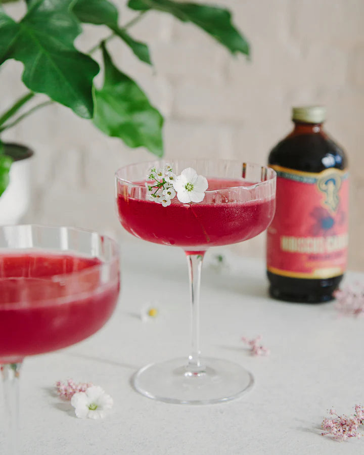 Cocktail in a coupe glass with a cardamom flower and the Hibiscus Cardamom syrup bottle on the table.