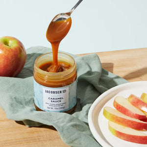 Someone pulling out a spoonful of Jacobsen salty caramel sauce from the jar next to a plate of apples.