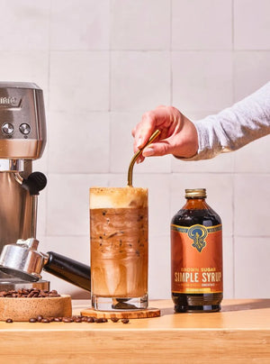 Person stirring their iced coffee drink with a straw, with the Brown Sugar Simple Syrup bottle on the counter.