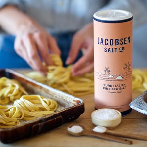 Cannister of Jacobsen Trapani Italian sea salt on a wood counter with a person making fresh pasta.